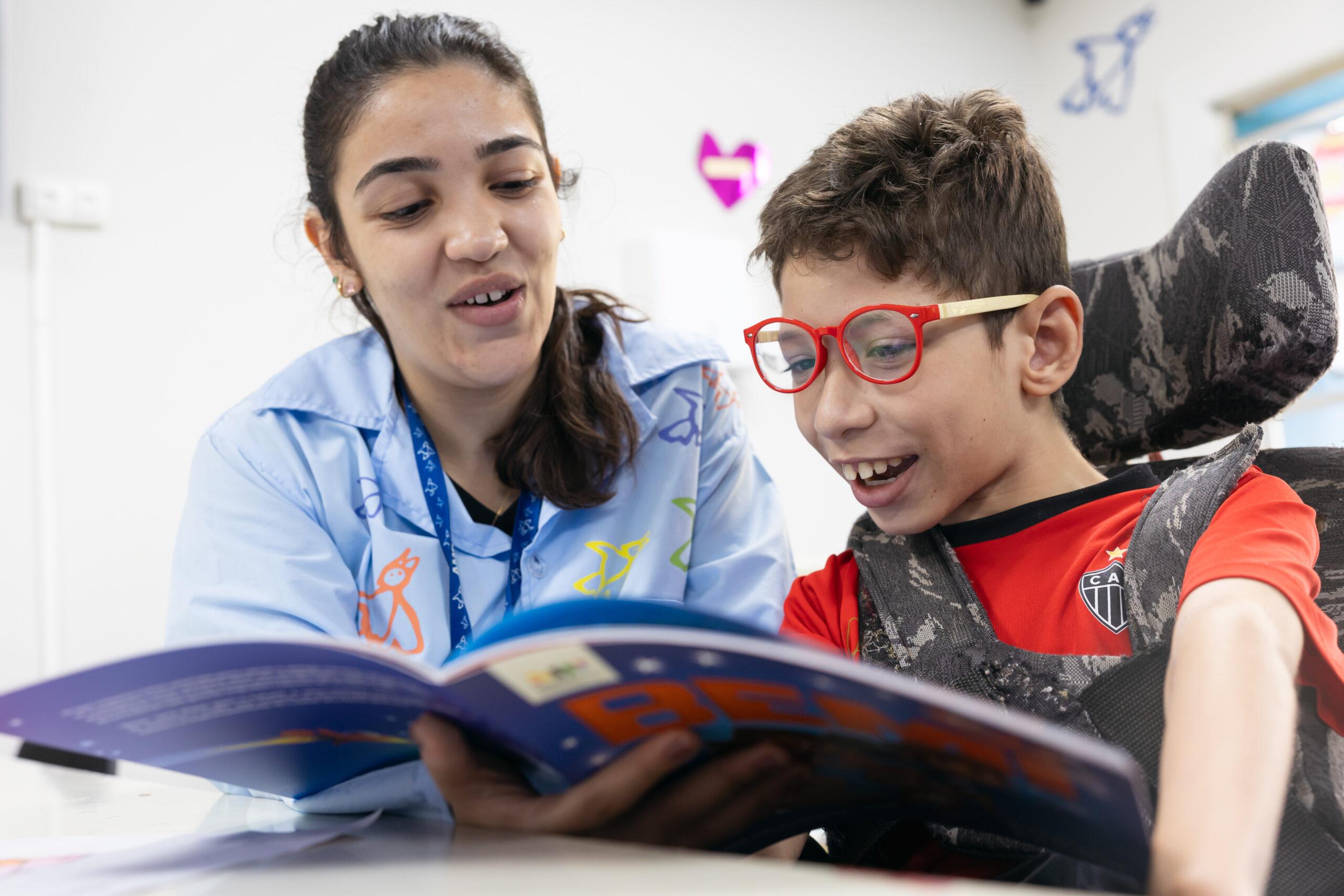 Criança com deficiência do projeto social AMR lendo um livro do Bemi - Uma chave para o futuro com o auxilio de uma professora