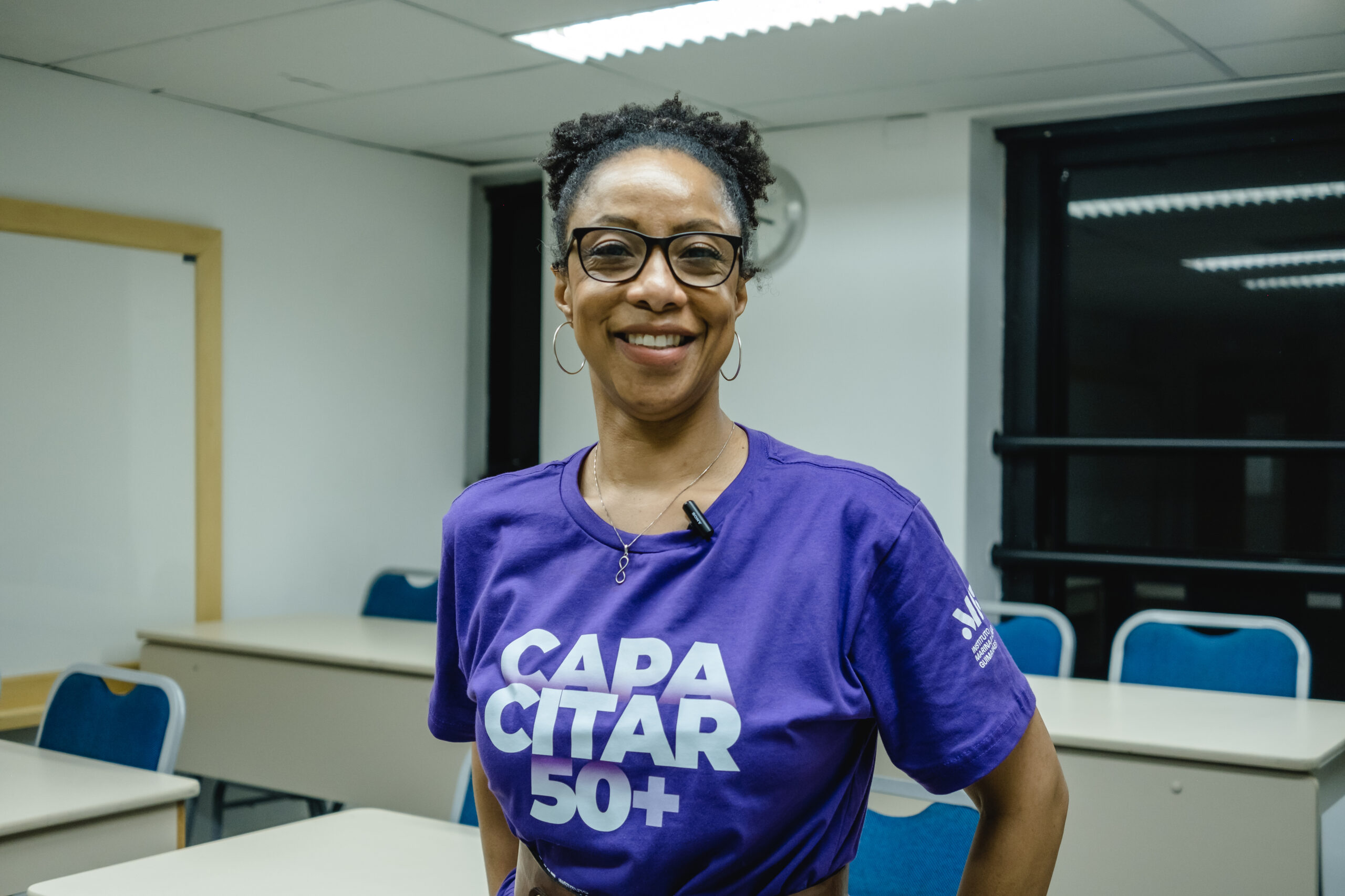 Aluna do projeto social Capacitar 50+ olhando e sorrindo para a foto com a sala de aula no fundo