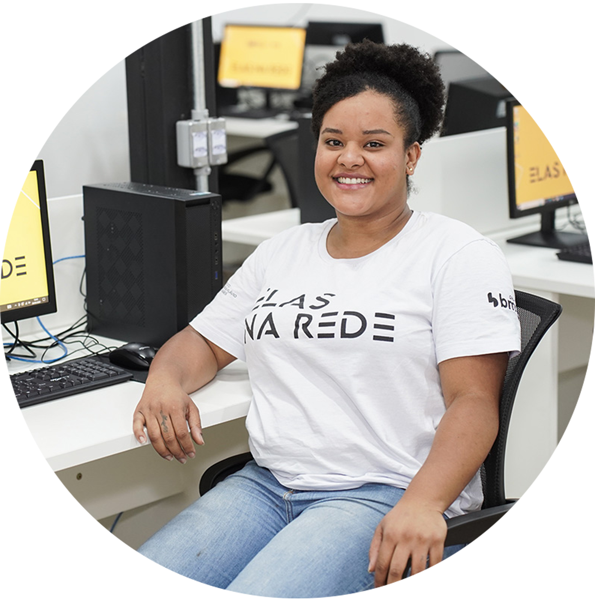 Aluna do projeto social Elas na Rede em sala de aula de frente e sorrindo para foto com computadores ao seu lado