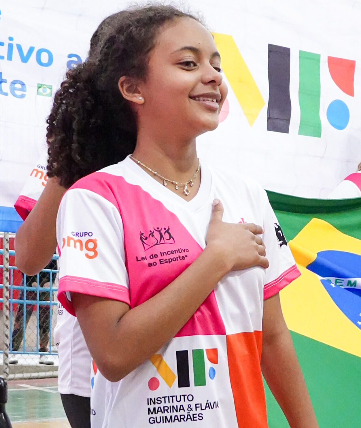 aluna de projeto social de voleibol cantando o hino nacional com a mão no peito e segurando a bandeira do Brasil